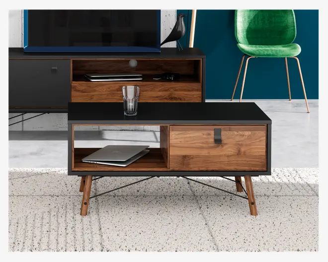 Rektangulært bord med sort topplade og træramme, udstyret med skrå træben og sorte metalstøtter. Bordet har en åben hylde til venstre med en lukket bærbar computer og et lukket rum med sort greb til højre. Et klart glas står ovenpå bordet.