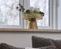 Guldfarvet fodstativ med en tekstureret, bølget kant, pyntet med et lille arrangement af mos, kogler og dekorative kugler ved et lyst vindue.