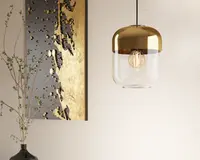 Loftlampe med et moderne design, der har en rund, metallisk gylden top og en klar glasbund. Pæren er synlig indeni, hvilket bidrager til det nutidige udtryk. Lampen hænger fra en sort ledning, og det samlede udseende er stilrent og elegant, velegnet til moderne indretning.