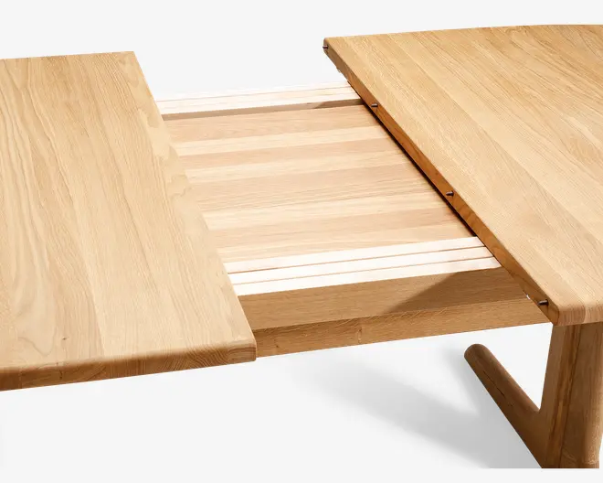 Nærbillede af et spisebord i træ med udtræksfunktion. Bordet er vist delvist åbent, så tillægspladen i midten er synlig. Træet har en lys, naturlig finish med tydelige åretegninger. Bordpladens kanter er let afrundede, og tillægspladen passer sømløst sammen med hovedbordet, når det er lukket.