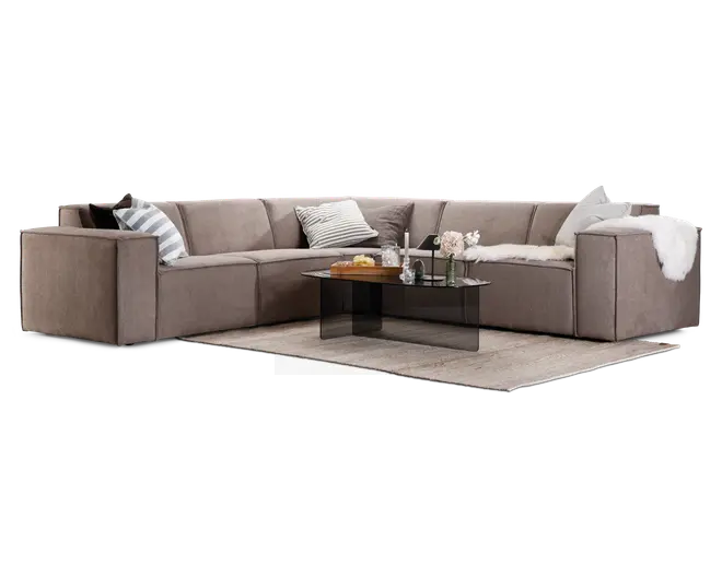 Stor L-formet sofa med brede, lave armlæn og blødt taupefarvet stofbetræk. Sæderne er dybe og komfortable, og der er flere pyntepuder i neutrale farver og striber. Et hvidt, kunstpels tæppe er lagt over det ene armlæn. Sofaen står på et lyst gulvtæppe og er kombineret med et moderne sofabord i sort glas, hvorpå der står stearinlys, blomster og en bakke med drikkevarer og snacks.