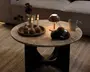 Rund beige marmorbord med tyk stenplade og sort skulpturel sokkel, stylet med en bakke med stearinlys og små vaser, en tændt bordlampe, garnnøgler og en keramikkop.
