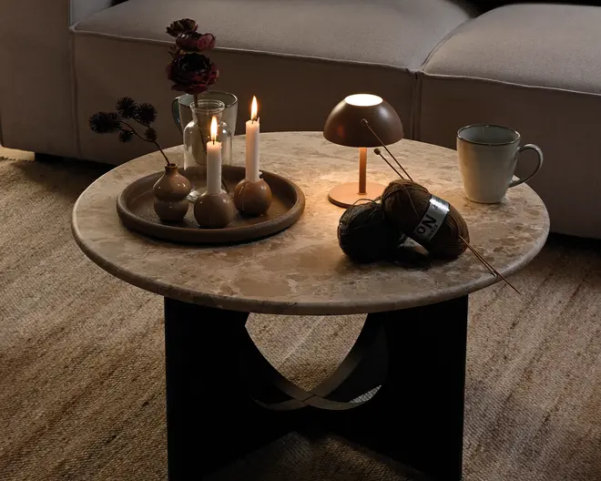 Rund beige marmorbord med tyk stenplade og sort skulpturel sokkel, stylet med en bakke med stearinlys og små vaser, en tændt bordlampe, garnnøgler og en keramikkop.