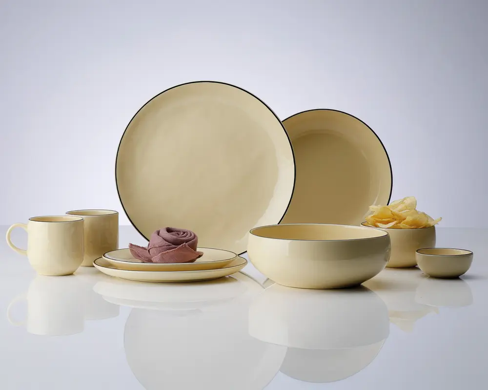 Et sæt cremefarvet keramikservice med blank overflade, bestående af to store tallerkener, en mellemstor skål, en lille skål, to krus og en sidetallerken. Delene har en diskret mørk kant og et glat, minimalistisk design. Sættet er arrangeret pænt, så man kan se de forskellige størrelser og former, der egner sig til servering af mad og drikke.