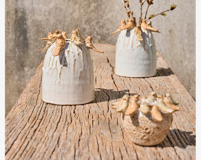 Tre håndlavede keramikvaser med rustik, tekstureret overflade står på en vejrbidt træflade. Hver vase er dekoreret med små, modellerede fugle, der sidder rundt om kanten og giver et finurligt og naturligt udtryk. Vaserne varierer i størrelse og form, hvor to har en glat, lys glasur, og én har en mere porøs, jordagtig tekstur. Fuglene er glaseret i en varm, lysebrun farve, som står i blid kontrast til vaserens neutrale nuancer.