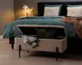 Rektangulær polstret opbevaringsbænk med hængslet låg, betrukket med et tekstureret beige stof. Bænken står på fire slanke sorte metalben og vises med låget åbent, hvilket afslører et indvendigt opbevaringsrum med puder og et sammenfoldet tæppe. Designet er moderne og funktionelt, velegnet til placering for enden af en seng.