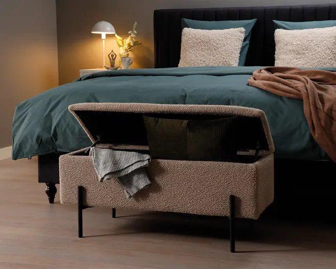 Rektangulær polstret opbevaringsbænk med hængslet låg, betrukket med et tekstureret beige stof. Bænken står på fire slanke sorte metalben og vises med låget åbent, hvilket afslører et indvendigt opbevaringsrum med puder og et sammenfoldet tæppe. Designet er moderne og funktionelt, velegnet til placering for enden af en seng.