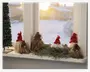 En gruppe små dekorative nissefigurer med lange skæg og røde huer er arrangeret på en vindueskarm. Nogle har hvide skæg, mens andre har brune, strå-lignende skæg. Nisserne er omgivet af kunstige grangrene, hvilket giver et festligt og vinterligt udtryk. Sollys strømmer ind gennem vinduet og skaber en hyggelig julestemning.