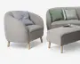 Billedet viser et sæt moderne møbler med lysegråt betræk, afrundede kanter og bløde konturer. Lænestolen har en buet ryg og armlæn, understøttet af fire lyse træben, og indeholder en blød, lyseblå pude. Sofaen har et lignende design med matchende betræk og to pyntepuder – én ensfarvet grøn og én stribet i grøn, blå og beige. Skamlen er rektangulær med let afrundede hjørner, også med lyse træben, og der ligger et magasin ovenpå. Stilen er minimalistisk og moderne med fokus på komfort og rene linjer.