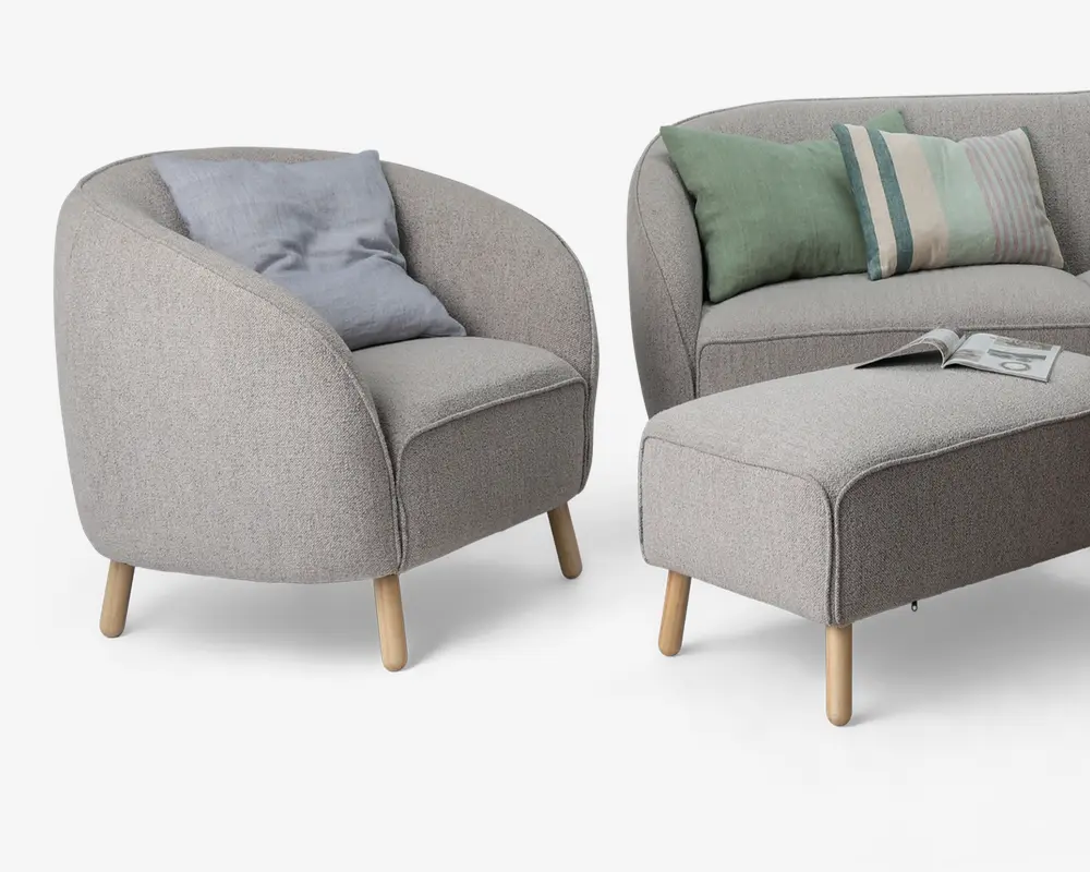 Billedet viser et sæt moderne møbler med lysegråt betræk, afrundede kanter og bløde konturer. Lænestolen har en buet ryg og armlæn, understøttet af fire lyse træben, og indeholder en blød, lyseblå pude. Sofaen har et lignende design med matchende betræk og to pyntepuder – én ensfarvet grøn og én stribet i grøn, blå og beige. Skamlen er rektangulær med let afrundede hjørner, også med lyse træben, og der ligger et magasin ovenpå. Stilen er minimalistisk og moderne med fokus på komfort og rene linjer.