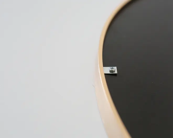Nærbillede af bagkanten på et rundt objekt med en lys kant og en mørk overflade. En lille metalbeslag med en skrue er fastgjort til kanten, sandsynligvis til ophængning eller fastgørelse af produktet.
