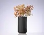Høj, mat sort cylindrisk vase med lodrette riller, fyldt med en løs buket af tørrede beige og rosa blomster og blade.