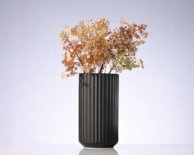 Høj, mat sort cylindrisk vase med lodrette riller, fyldt med en løs buket af tørrede beige og rosa blomster og blade.