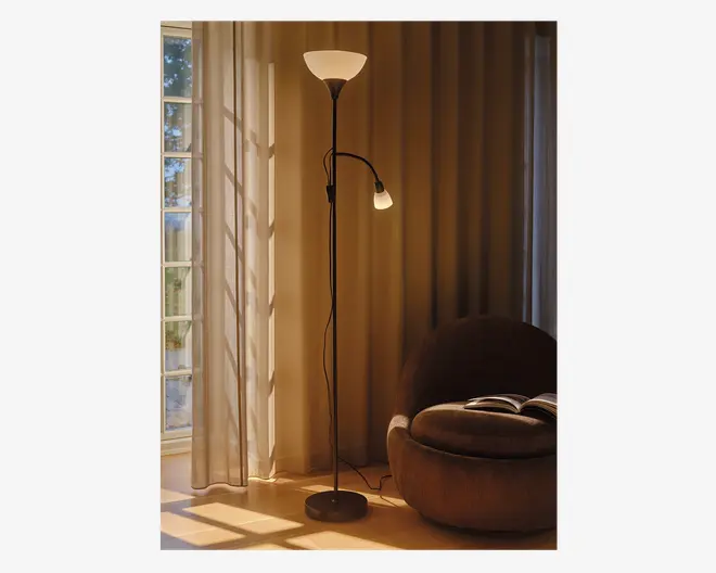 Høj, sort gulvlampe med to lyskilder: en opadrettet, skålformet hovedlampe og en mindre, justerbar læselampe fastgjort til stangen. Lampen har en rund, stabil fod og et enkelt, moderne design. Læselampen kan justeres efter behov, hvilket gør lampen velegnet til både stemnings- og arbejdsbelysning. Lampen står ved siden af et stort vindue med lette gardiner og en hyggelig, rund lænestol med en åben bog på.