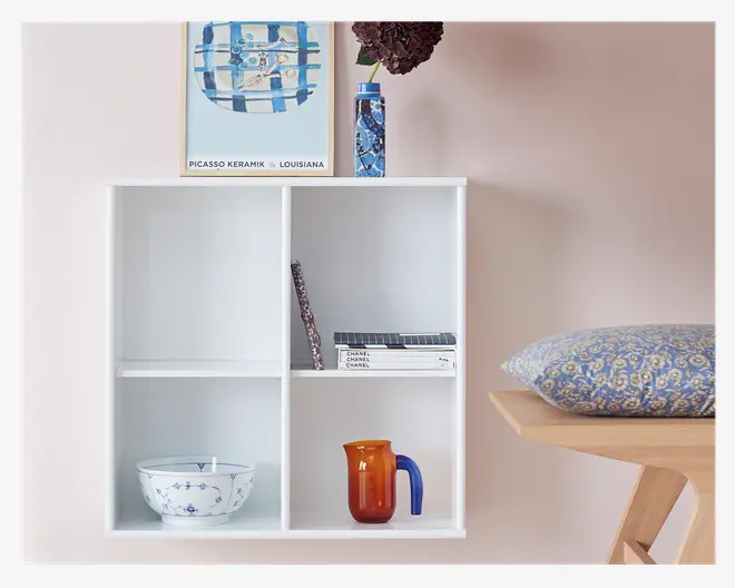 En hvid væghylde med fire rum ses mod en lyserød væg. Øverste venstre rum er tomt, mens nederste venstre rum indeholder en hvid keramikskål med blå blomsterdekoration. Øverste højre rum har nogle stablede bøger og en dekorativ genstand, og nederste højre rum rummer en lille ravfarvet glaskande med blåt håndtag. Oven på hylden står et indrammet abstrakt kunsttryk i blå nuancer, en høj vase med blåt mønster og mørkerøde blomster, og til højre ses en del af en træbænk med en blå og gul blomsterpude.