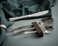 Et sæt blankt bestik i rustfrit stål med et klassisk, let dekoreret skaftdesign. Sættet består af en middagskniv, en gaffel, en spiseske og en teske. Bestikket er placeret på en blød, blågrøn tekstil, hvor tre dele hviler på en brun bestikholder af filt, som løfter dem fra bordfladen. Bestikket har en reflekterende overflade og et traditionelt, elegant udseende.