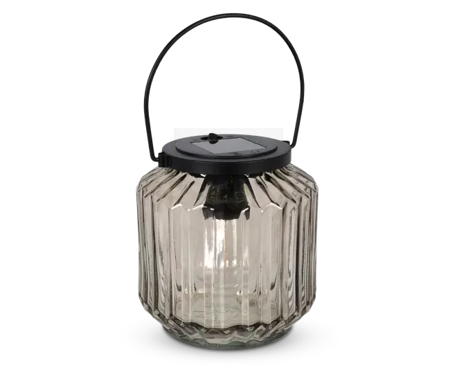 Lanterne af klart glas med lodret rillet struktur, sort metal låg med indbygget solcellepanel og matchende sort metalhank til ophængning eller transport. Lanternen har en rund, let facetteret form og er designet til udendørs brug, hvor den giver stemningsfuld belysning.