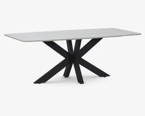 Rektangulært bord med en glat, lys bordplade og afrundede kanter. Understellet har et markant, moderne design med sorte, skrå metalben, der krydser hinanden i midten og danner et stjerne- eller edderkoppelignende udtryk. Bordet har et minimalistisk og nutidigt look, velegnet til spise- eller kontorbrug.