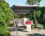 Udendørs gyngesofa med solid metalramme og gråt soltag. Sædet har et lameldesign og er pyntet med to dekorative puder med blomstermønster, en rund rød pude og et lyserødt tæppe, der er lagt over ryglænet. Gyngen står på et grusunderlag og er omgivet af frodig beplantning.
