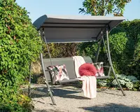 Udendørs gyngesofa med solid metalramme og gråt soltag. Sædet har et lameldesign og er pyntet med to dekorative puder med blomstermønster, en rund rød pude og et lyserødt tæppe, der er lagt over ryglænet. Gyngen står på et grusunderlag og er omgivet af frodig beplantning.