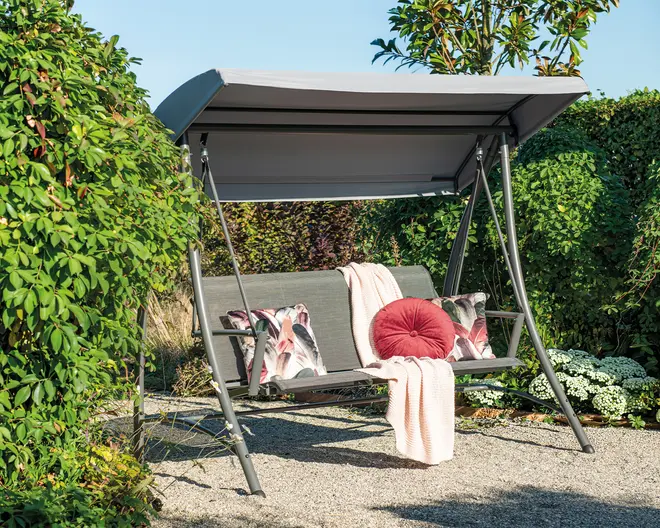 Udendørs gyngesofa med solid metalramme og gråt soltag. Sædet har et lameldesign og er pyntet med to dekorative puder med blomstermønster, en rund rød pude og et lyserødt tæppe, der er lagt over ryglænet. Gyngen står på et grusunderlag og er omgivet af frodig beplantning.