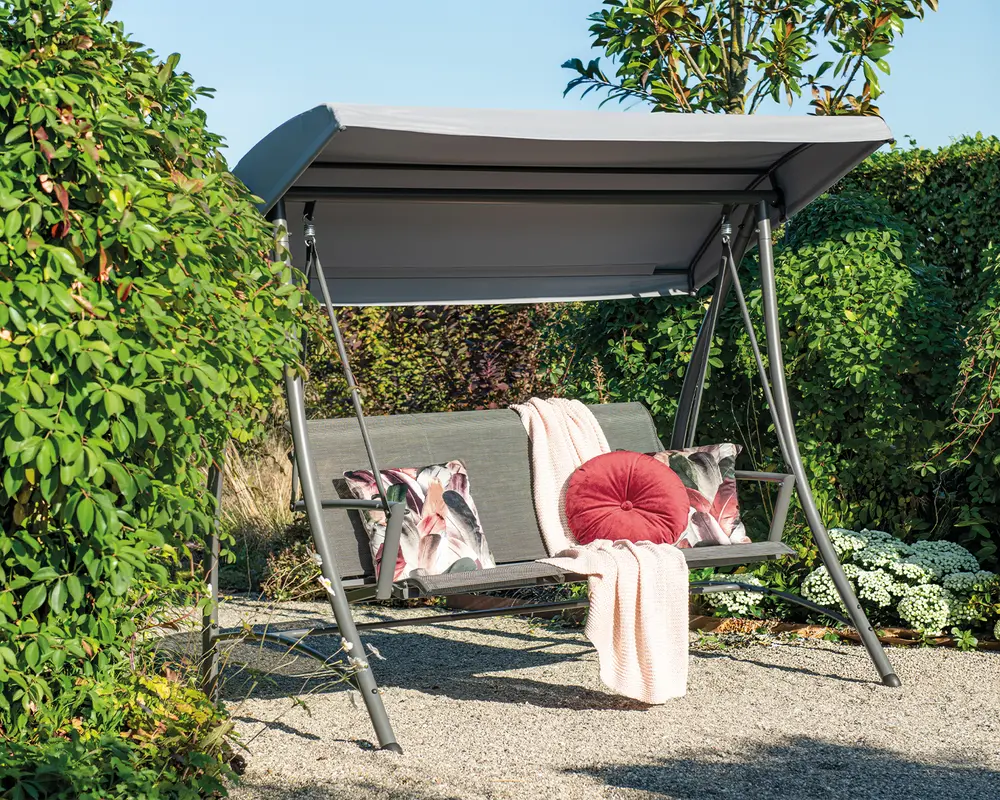 Udendørs gyngesofa med solid metalramme og gråt soltag. Sædet har et lameldesign og er pyntet med to dekorative puder med blomstermønster, en rund rød pude og et lyserødt tæppe, der er lagt over ryglænet. Gyngen står på et grusunderlag og er omgivet af frodig beplantning.