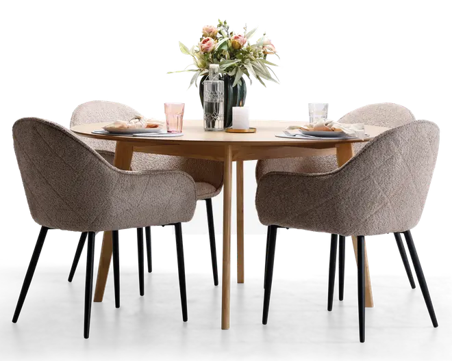 Et rundt spisebord i lyst træ med fire polstrede stole i blødt beige stof med quiltet struktur og sorte, tilspidsede ben. Bordet er dækket op til fire personer med tallerkener, servietter, glas og bestik. I midten af bordet står en dekorativ opstilling med en vase med friske blomster, en glasflaske og et stearinlys, hvilket skaber en indbydende og elegant spiseatmosfære.