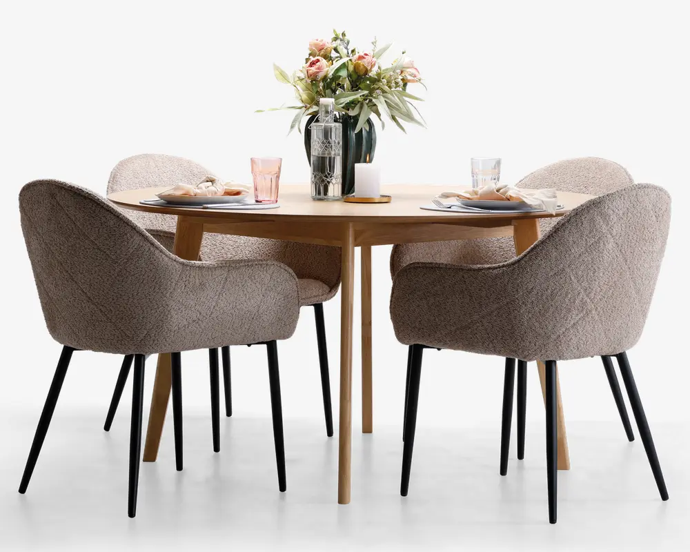 Et rundt spisebord i lyst træ med fire polstrede stole i blødt beige stof med quiltet struktur og sorte, tilspidsede ben. Bordet er dækket op til fire personer med tallerkener, servietter, glas og bestik. I midten af bordet står en dekorativ opstilling med en vase med friske blomster, en glasflaske og et stearinlys, hvilket skaber en indbydende og elegant spiseatmosfære.