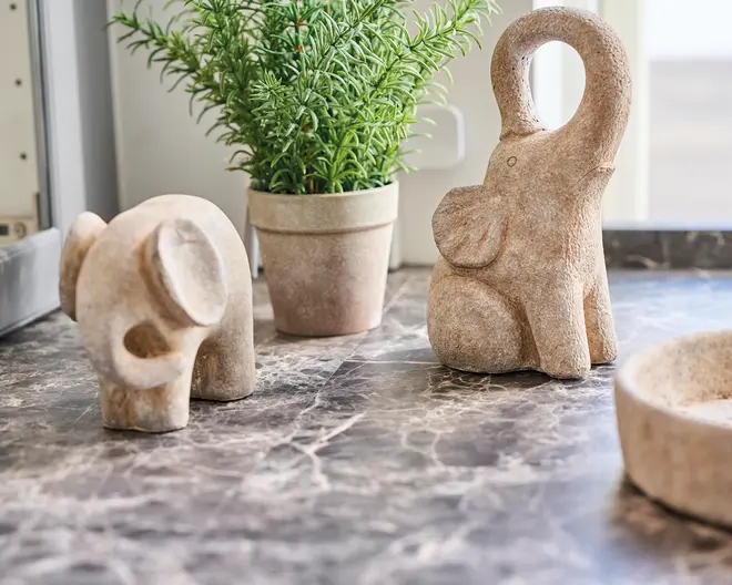 To dekorative elefantfigurer lavet af lyse sten står på en bordplade med marmormønster. Den ene elefant sidder med snablen løftet i en cirkelform, mens den anden bøjer sig fremad med snablen nedad. Begge figurer har et glat, minimalistisk design med diskrete udskårne detaljer. I baggrunden står en grøn potteplante i en enkel beige potte, som tilfører et naturligt præg til opstillingen.