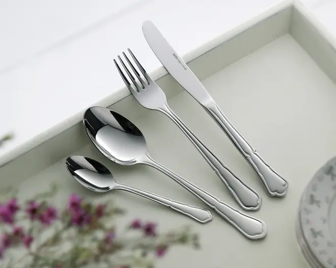 Fire stykker blankt bestik i rustfrit stål ligger på en lys bakke: en middagskniv, middagsgaffel, spiseske og teske. Hvert stykke har et klassisk, let dekorativt håndtag med bløde kurver og en udsmykket kant nær enden. Overfladerne er meget blanke, og stilen er elegant og egnet til både hverdag og fest.
