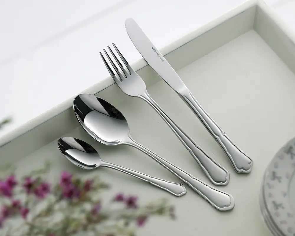 Fire stykker blankt bestik i rustfrit stål ligger på en lys bakke: en middagskniv, middagsgaffel, spiseske og teske. Hvert stykke har et klassisk, let dekorativt håndtag med bløde kurver og en udsmykket kant nær enden. Overfladerne er meget blanke, og stilen er elegant og egnet til både hverdag og fest.