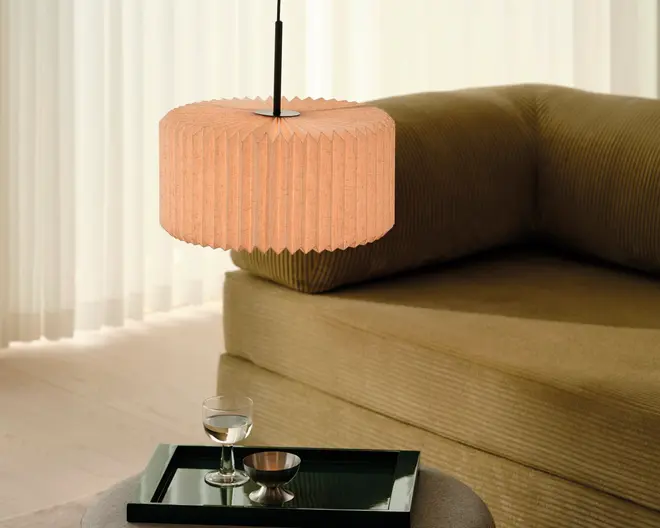 Ferskenfarvet, plisseret cylindrisk pendelskærm med sort ledning, vist hængende over en moderne sofa i et lyst, minimalistisk rum.