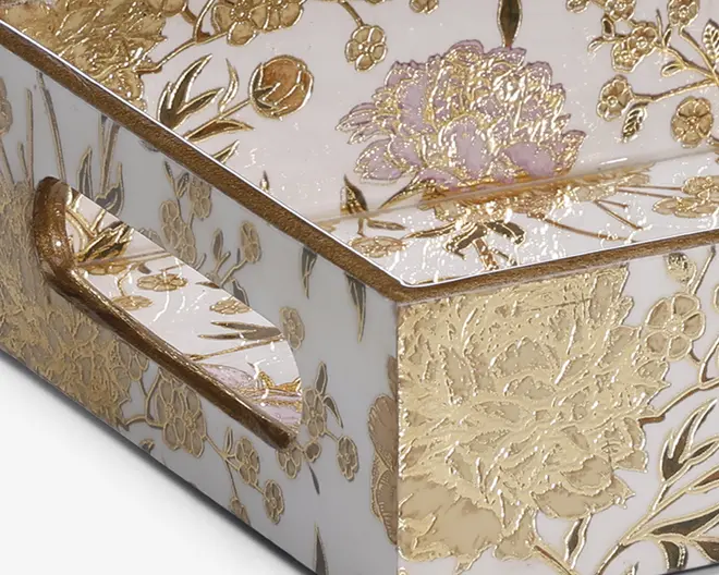 Nærbillede af et hjørne af en æske med en blank hvid overflade, dekoreret med indviklede guldfarvede blomster og detaljer. Blomsterne er præget, hvilket giver en tekstureret, hævet effekt, og nogle kronblade har et diskret lavendelfarvet skær. Æsken har et matchende håndtag med samme guldfarvede blomstermønster, og det samlede udtryk er luksuriøst og dekorativt.