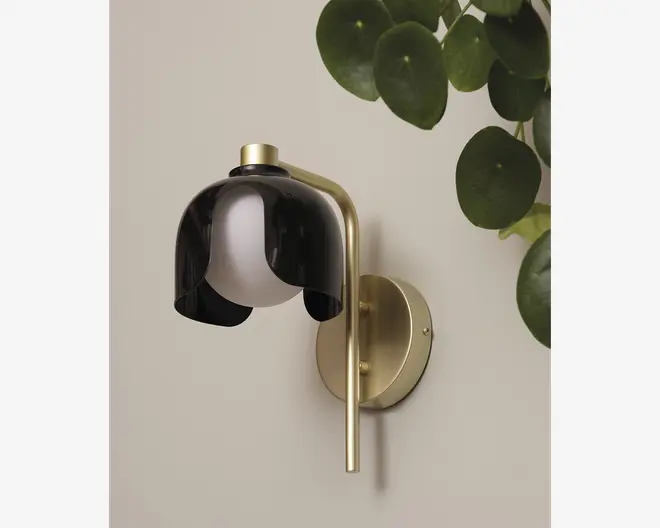Væghængt lampe med rund, børstet messingbase og arm. Lampen har en kuppelformet sort skærm med blank finish, der er delvist åben i bunden og afslører en hvid kugleformet pære. Designet er moderne og elegant med en kombination af metal og mørke nuancer.