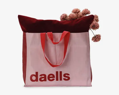 Stor genanvendelig indkøbspose lavet af lyserødt stof med røde sidepaneler og håndtag. Forsiden har teksten 'daells' i store, røde bogstaver. Posen er vist fyldt med en mørkerød velourpude og kunstige lyserøde pompon-blomster, hvilket viser dens rummelige kapacitet og solide konstruktion.