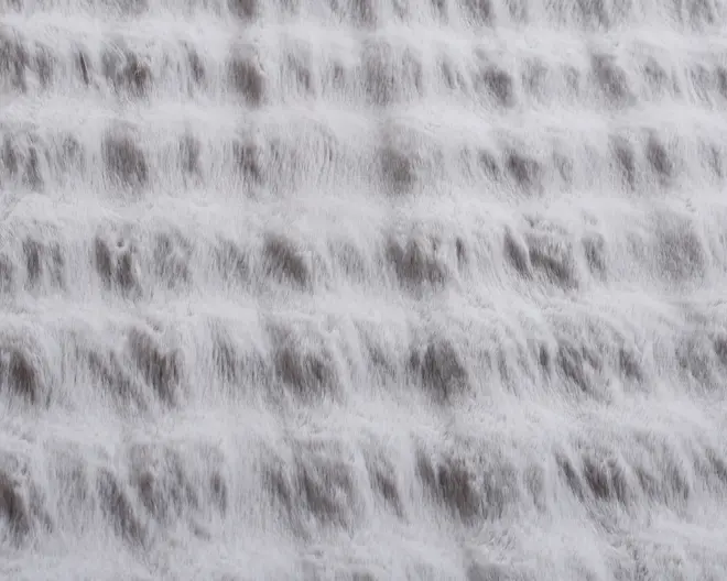 Nærbillede af rindende vand, der strømmer ned over flere vandrette trin og danner et tekstureret, lagdelt udtryk med bløde, hvide skummønstre og diskrete skygger.