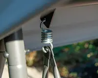 Nærbillede af en solid metalfjeder-mekanisme fastgjort til en ramme, hvor en spiralformet stålfjeder er forbundet med en krog og to metalsupportstænger. Delene ser ud til at være en del af en udendørs konstruktion med mat grå finish og robust design for stabilitet og holdbarhed.