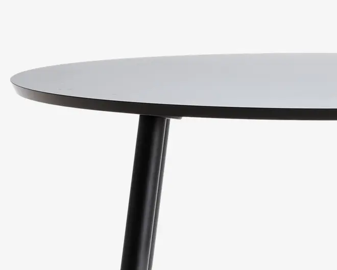Nærbillede af en rund bordplade med en glat, lysefarvet overflade og en tynd, mørk kant. Bordet har et robust, cylindrisk sort ben fastgjort under, hvilket giver det et moderne og minimalistisk udseende.