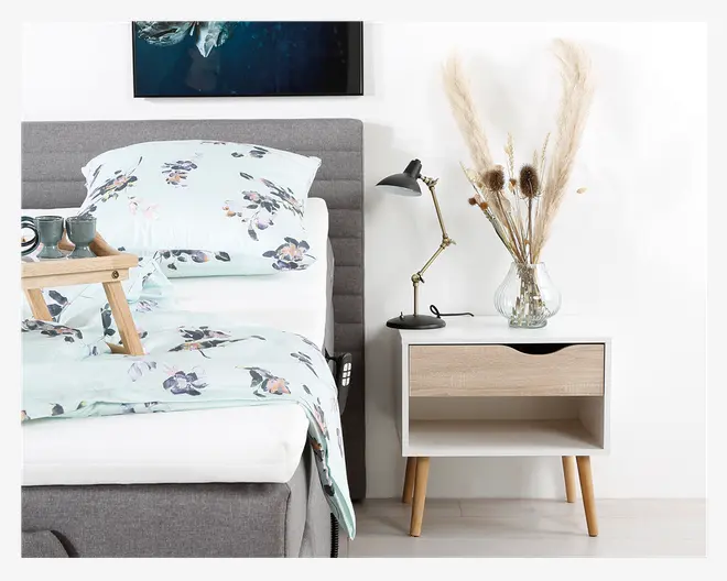 Et moderne natbord med hvid ramme, front i lyst træ og skrå træben. Bordet har en enkelt skuffe med sort halvcirkelformet udskæring som greb og en åben hylde nedenunder. Ovenpå natbordet står en klar glasvase med høje, tørrede græsser og blomster samt en sort, justerbar læselampe med messingarm.