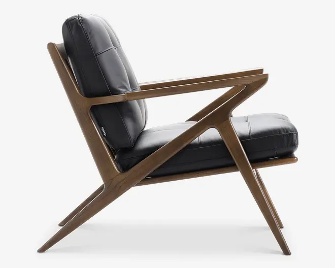 Sidevisning af en moderne loungestol med en slank, vinklet træramme i naturlig finish. Stolen har tykke, sorte læderhynder på sæde og ryglæn, som giver komfort og støtte. Armlænene er integreret i den geometriske ramme, og benene er skråtstillede for stabilitet. Det samlede design kombinerer moderne stil med inspiration fra midten af det 20. århundrede.