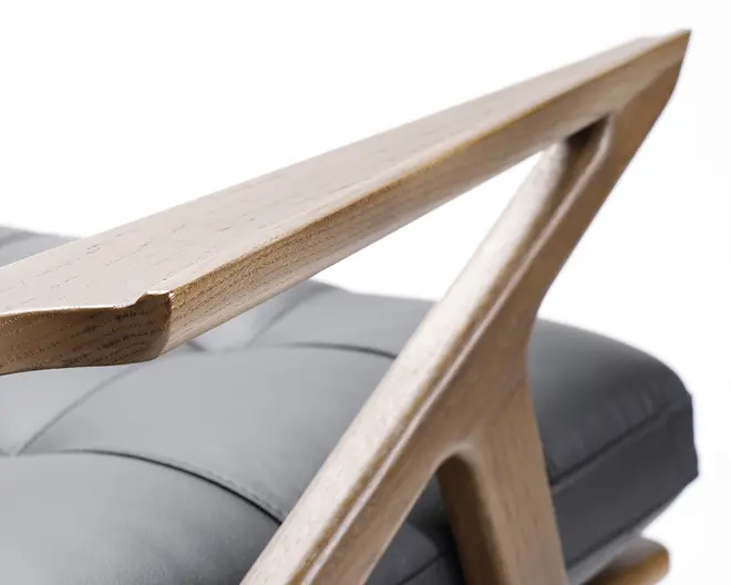 Nærbillede af et stolearmlæn lavet af lyst træ med et glat, vinklet design. Armlænet har en geometrisk, trekantet støtte. Sædepuden er polstret med blødt, gråt læder og har synlige syninger, hvilket giver et moderne og komfortabelt udtryk.