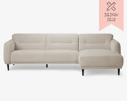 Moderne L-formet sofa betrukket med et lyst beige stof med en blød, tekstureret overflade. Sofaen har tre sædehynder og tre ryghynder med diskrete, horisontale syninger. Højre side har en chaiselong for ekstra komfort. Sofaen har slanke, let skrå armlæn og står på lave, sorte, cylindriske ben, hvilket giver et moderne og minimalistisk udtryk.