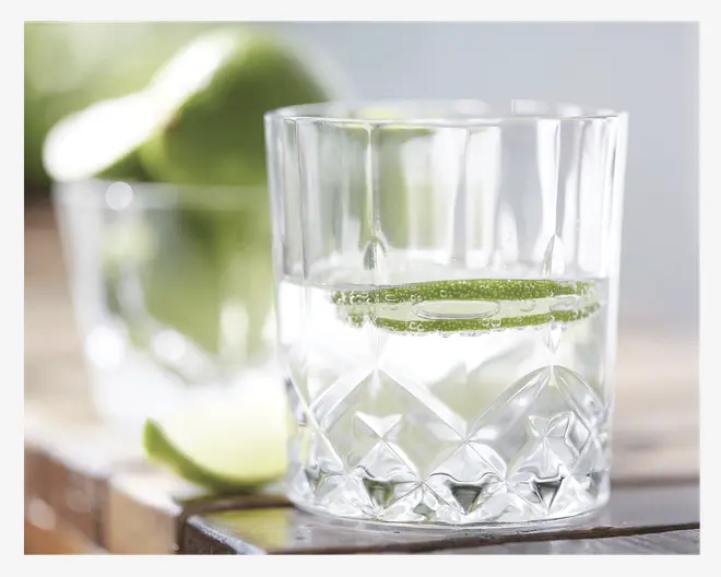 Et klart glas med facetslebet, geometrisk diamantmønster i bunden, fyldt med en gennemsigtig væske og en skive lime. Glasset har en tyk, solid bund og lige sider. I baggrunden ses et andet glas samt hele og skivede limefrugter, let ude af fokus.