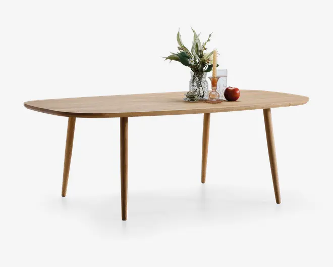 Rektangulært bord i lyst egetræ med glat overflade og afrundede hjørner. Bordet har fire koniske ben og et minimalistisk design. Ovenpå står en klar glasvase med grønne grene, et højt stearinlys, en lille dekorativ flaske, et rødt æble og en bog, hvilket giver et naturligt og hyggeligt udtryk.