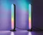 Et par slanke, lodrette LED-lysstænger med afrundede kanter, monteret på cirkulære sorte baser. Lysene viser en farverig gradient med blå, grøn, gul og pink, som kaster et farverigt skær på væggen og overfladen. Hver lysstang er forbundet med en synlig sort ledning. Designet er moderne og minimalistisk, velegnet til stemnings- eller accentbelysning.