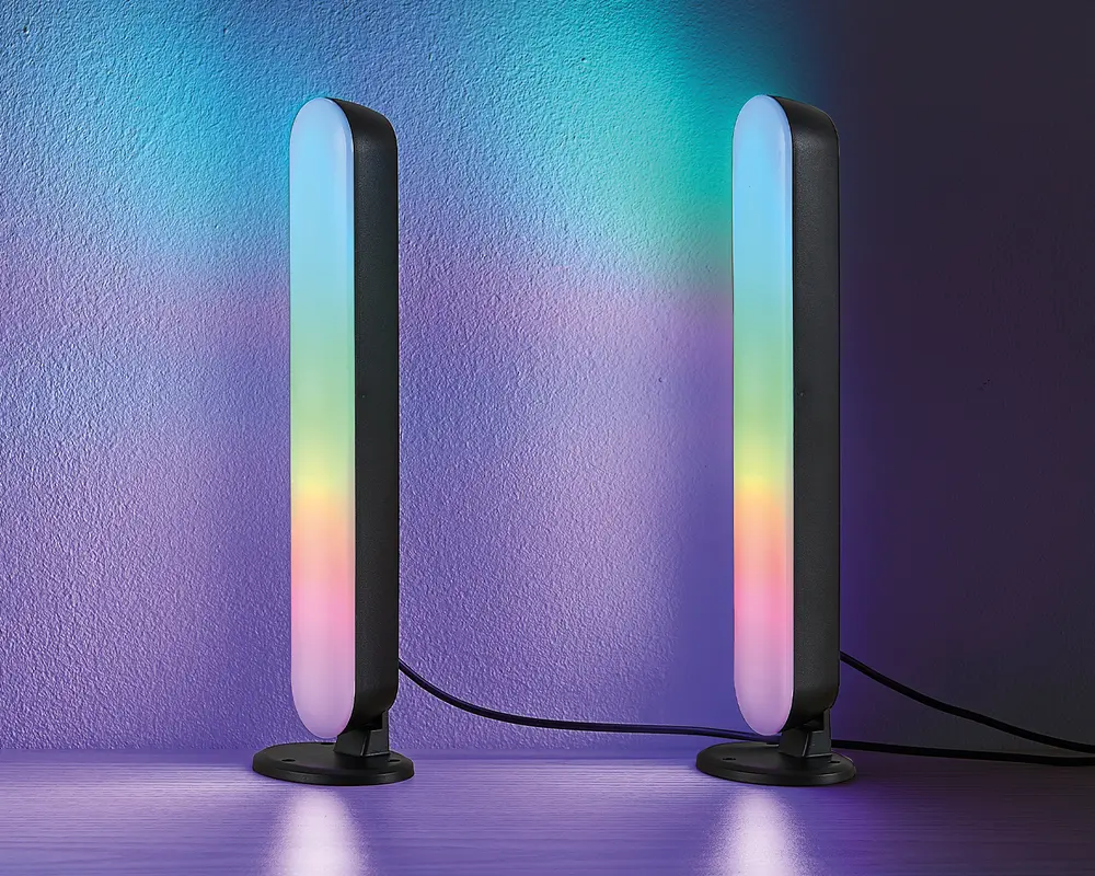 Et par slanke, lodrette LED-lysstænger med afrundede kanter, monteret på cirkulære sorte baser. Lysene viser en farverig gradient med blå, grøn, gul og pink, som kaster et farverigt skær på væggen og overfladen. Hver lysstang er forbundet med en synlig sort ledning. Designet er moderne og minimalistisk, velegnet til stemnings- eller accentbelysning.