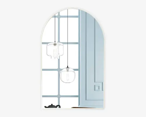 Billedet viser et dekorativt vægspejl med buet top. Spejlet har et enkelt, rammeløst design og reflekterer et moderne interiør med to pendellamper med klare glasskærme, en lyseblå væg med paneler og et vindue med gittermønster. En sort tekande kan ses på en overflade i nederste venstre hjørne af spejlbilledet.
