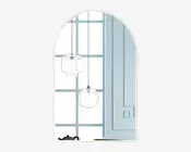 Billedet viser et dekorativt vægspejl med buet top. Spejlet har et enkelt, rammeløst design og reflekterer et moderne interiør med to pendellamper med klare glasskærme, en lyseblå væg med paneler og et vindue med gittermønster. En sort tekande kan ses på en overflade i nederste venstre hjørne af spejlbilledet.