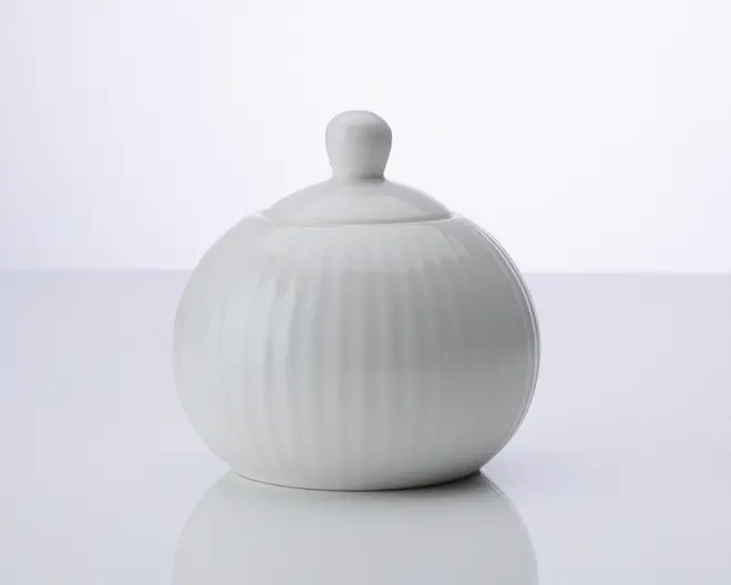 Rund, hvid keramisk beholder med tilhørende låg, der har et knopformet håndtag. Beholderen har diskrete lodrette riller, som giver tekstur, og det samlede design er glat og minimalistisk.