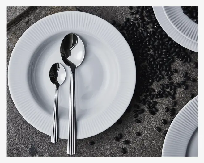 To rustfri stålskeer, en stor og en lille, med et moderne design og lodrette riller på skaftet, ligger på en hvid keramik-tallerken med diskret rillet kant. Tallerkenen står på en grå, tekstureret overflade, hvor der er spredte sorte bønner og delvist synlige lignende tallerkener i billedets kanter.